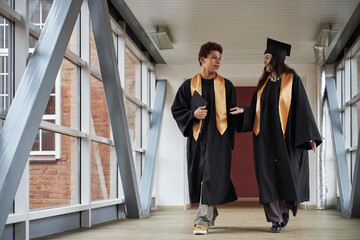 Young Caucasian man and woman wearing graduation gowns walking together in university corridor,...