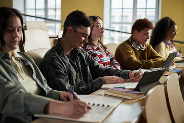 Group of diverse university students sitting in lecture hall taking notes and using digital tablets, young people including Caucasian and Black individuals focusing on academic work during class