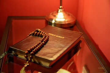 Holy Bible with prayer beads on table near red wall