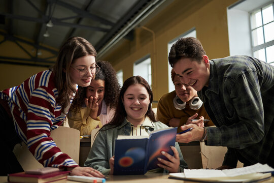 Group of multiethnic university students gathering around desk smiling and discussing textbook together, young adults collaborating on academic project in classroom setting