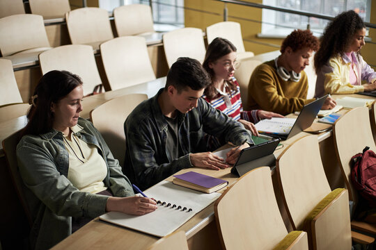 Group of multiethnic young students sitting in lecture hall taking notes and using digital devices during university class, focusing on studying and academic work