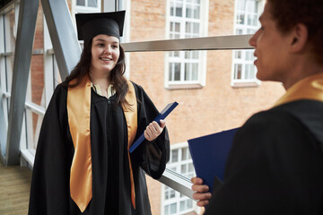 Caucasian young woman, wearing graduation cap and gown, smiling while holding diploma folder and...