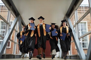 Group of multiethnic young university students wearing graduation caps and gowns running together...