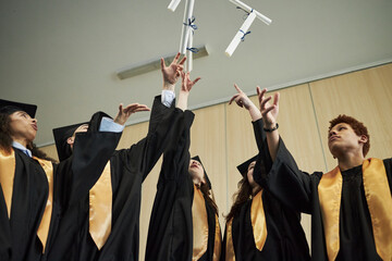 Group of diverse university young students wearing graduation gowns celebrating by tossing diplomas...