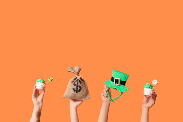 Female hands with tasty cupcakes, leprechaun hat and money bag for St. Patrick's Day on orange...