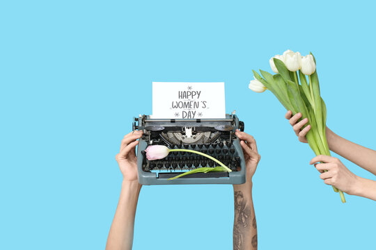 Hands holding vintage typewriter, tulips and festive postcard with text HAPPY WOMEN'S DAY on blue background