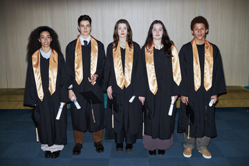 Group of five multiethnic teenagers standing in graduation gowns holding diplomas, facing camera...