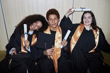 Diverse group of cheerful young students wearing graduation gowns sitting together smiling and...