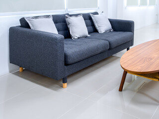 Empty grey cozy fabric sofa seat with pillows near oval wooden table on clean tiles floor, near roller blinds at the window, wait area on white modern living room with nobody.