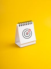 Target dart bullseye icon on white desk calendar page isolated on yellow background, vertical photo. Business goal and success, scheduled, agenda, action plan concepts in new year.