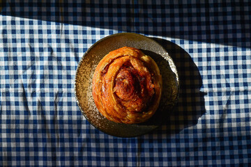 Top view of Danish roll on the plate with sun light, shadows isolated on napkin background. Dark, black, pastry, sweet bun, sugar, bread. High angle, above, close up.