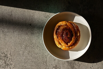 Top view of Danish roll on the plate with sun light and shadows. Dark, black, pastry, sweet bun, sugar, bread. Grey or gray cement on the background. High angle, above. Copy space, empty, free.