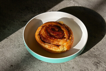 Close up view of Danish roll on the plate with sun light and shadows. Dark, black, pastry, sweet bun, sugar, bread. Grey or gray cement on the background.