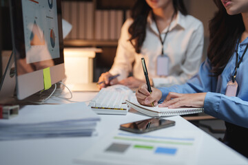 Businesswomen working late in office taking notes and analyzing charts on computer screen