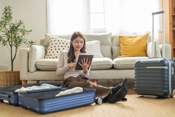 Asian woman traveler checking list on tablet preparing luggage packing clothes in suitcases for...