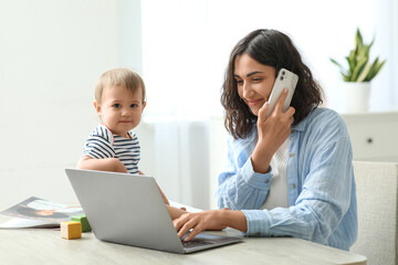 Cute baby and young mother working with laptop while talking by mobile phone on maternity leave at...