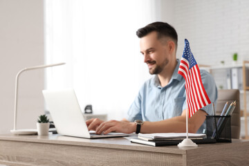Handsome young happy man with laptop and flag of USA working at table in office