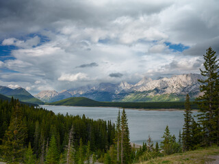 Wide scenic view of a mountain lake, dense forest, and rugged peaks under dramatic clouds in Peter Lougheed Provincial Park, Alberta. Classic Canadian wilderness in summer.