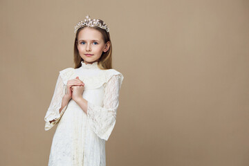 Young girl in elegant white dress and crown, showcasing a serene expression against a soft beige...