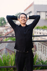 Smiling man in black clothing poses with hands behind head on a balcony with a good view