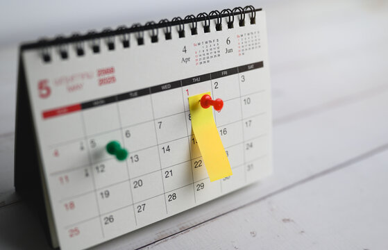 Close-up of desk calendar showing month of May 2025, Red and green push pins mark important dates, with yellow sticky note pinned on May 1st, symbolizing reminder or event planning