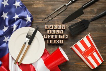 USA flag, wooden cubes with text HAPPY JULY FOURTH BBQ, dinnerware and barbecue utensils on wooden...