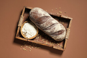 Tray with loaf of fresh rye bread, wheat and flour on brown background