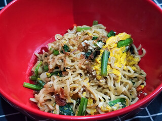A bright red bowl filled with delicious fried noodles with sliced ​​egg, mustard greens, crispy...