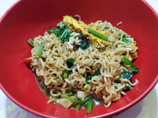 A bright red bowl filled with delicious fried noodles with sliced ​​egg, mustard greens, crispy fried onions and sliced ​​garlic, served on a tablecloth.