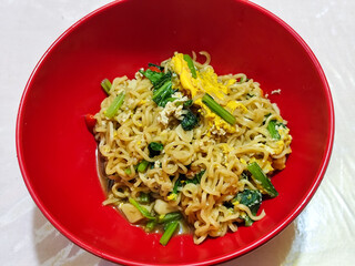 A bright red bowl filled with delicious fried noodles with sliced ​​egg, mustard greens, crispy...