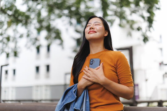 Young woman holding smartphone to chest, expressing gratitude and mindfulness outdoors
