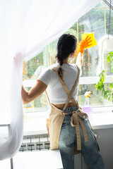 Woman in rubber gloves and apron manually washes window of house with rag cleaner and mop inside interior with home plants on windowsill. Restoring order and cleanliness in spring, cleaning servise