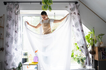 Woman in apron hangs transparent tulle curtains on large attic windows in the house inside the...