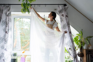 Woman in apron hangs transparent tulle curtains on large attic windows in the house inside the...
