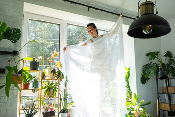 Woman in apron hangs transparent tulle curtains on large windows in the house inside the interior with potted plants. Spring cleaning, tidying up