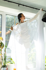 Woman in apron hangs transparent tulle curtains on large windows in the house inside the interior with potted plants. Spring cleaning, tidying up
