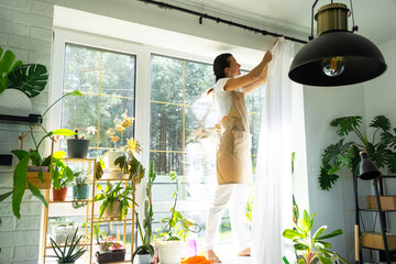 Woman in apron hangs transparent tulle curtains on large windows in the house inside the interior...