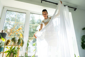 Woman in apron hangs transparent tulle curtains on large windows in the house inside the interior with potted plants. Spring cleaning, tidying up