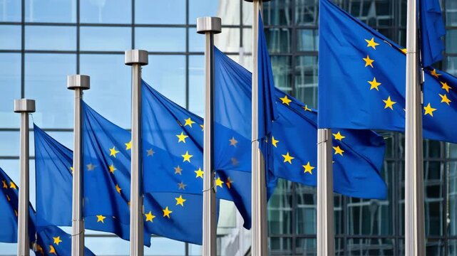 Multiple blue European Union flags flutter in the wind outside a contemporary building. The flags display golden stars against a rich blue backdrop under a clear sky