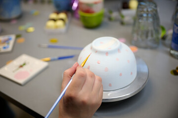 Artist Hand Painting Pink Polka Dots on Upside-Down Ceramic Bowl Using Fine Brush in a Creative Studio Setting with Art Supplies and Snacks