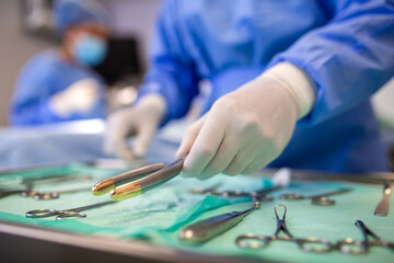 Close-up of a gloved hand picking up a surgical instrument from a sterile tray. Various surgical tools arranged on a green cloth in the operating room, with medical staff blurred in the background.