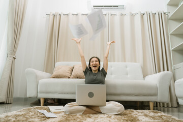 Asian woman throws papers in the living room as she finishes her work with her laptop on her lap.