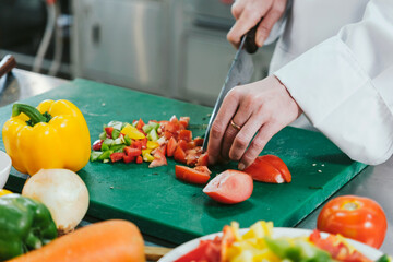 Chef in white uniform slicing fresh tomatoes on a green cutting board with chopped bell peppers in...