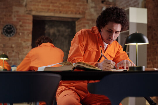 Young adult man wearing orange prison uniform sitting at table in prison library studying and writing in notebook with open books while another inmate sits in background