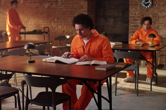 Young adult Caucasian man reading book at table in prison library, two other young adult men in orange uniforms studying in background, brick walls and books visible, educational setting