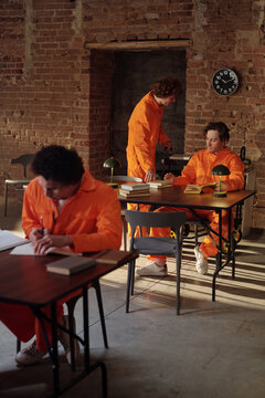 Three young adult men wearing orange prison uniforms studying and writing at desks in prison library, books and lamps on tables, exposed brick walls and clock in background