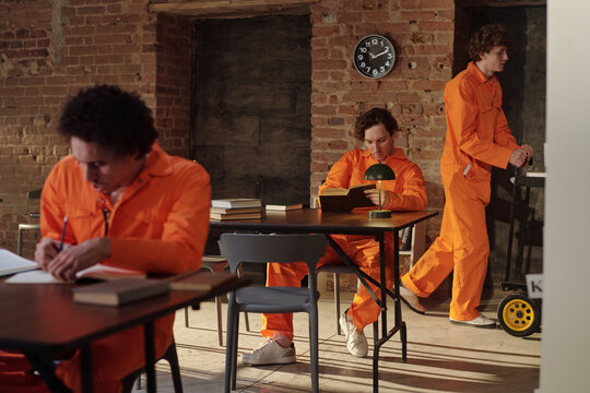 Three young men wearing orange uniforms studying and reading books in prison library, one sitting and writing at desk, another reading, third pushing cart with books