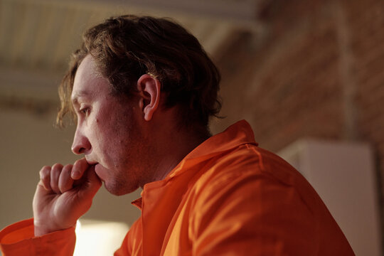 Caucasian young adult man sitting in prison library resting chin on hand appearing deep in thought, wearing orange uniform, brick wall and shelving visible in background