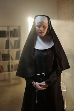 Caucasian woman dressed as nun standing in prison library holding closed Bible with rosary beads in hands, looking down with serious expression, bookshelves in background