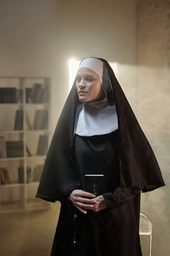Caucasian woman dressed as nun standing in prison library holding book with cross on cover, looking away from camera with serious expression, bookshelves in background